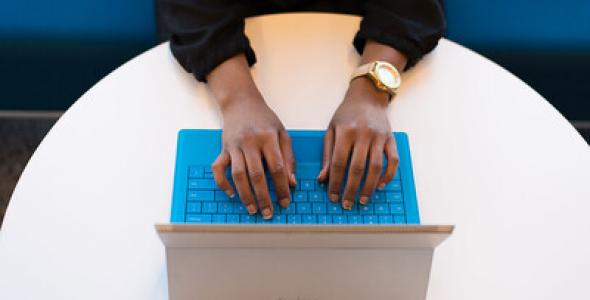 An aerial view of a woman's hands as she types at her laptop at a circular office table 