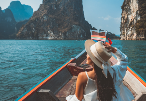 A woman sits in a boat in an exotic location, holding her hat.