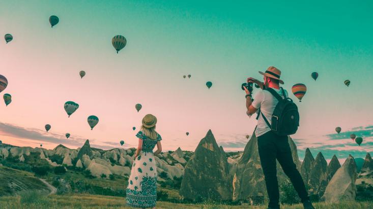 A man photographs a woman looking at a sky full of hot air balloons